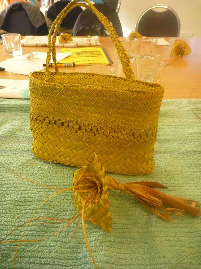 A finely woven golden pīngao kete (basket) with braided handles, displayed on a green surface at a workshop table, with a pīngao seed head in the foreground.