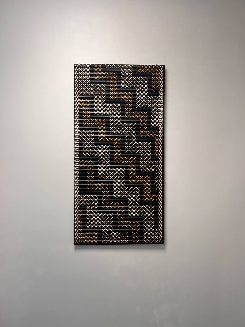 A rectangular tukutuku panel mounted on a white wall, featuring a traditional lattice weaving pattern in black, white, and golden pīngao.